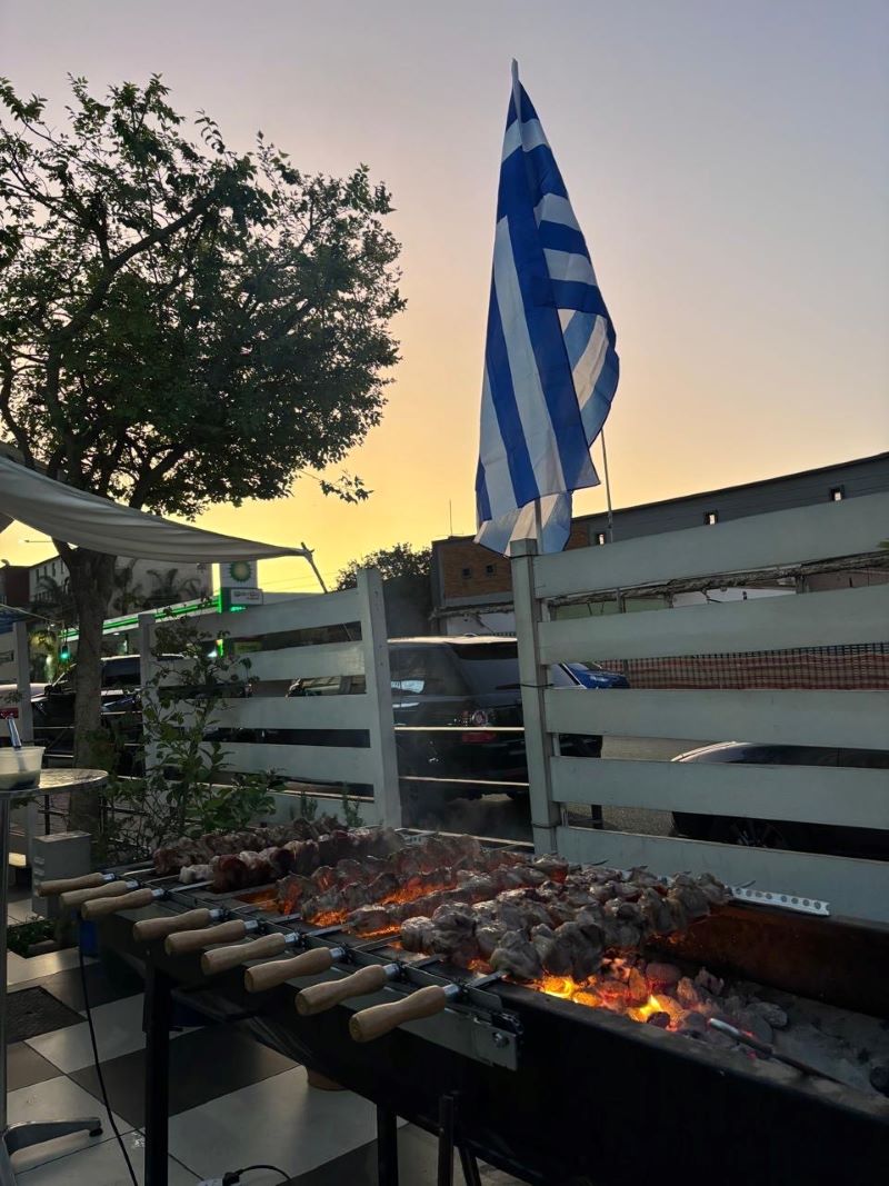 Souvlaki skewers grilling under the Greek flag at sunset